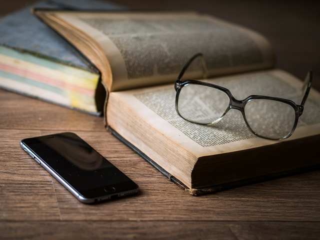 An open book is lying on a table with a pair of glasses on top. There is a mobile phone on the table next to it.
