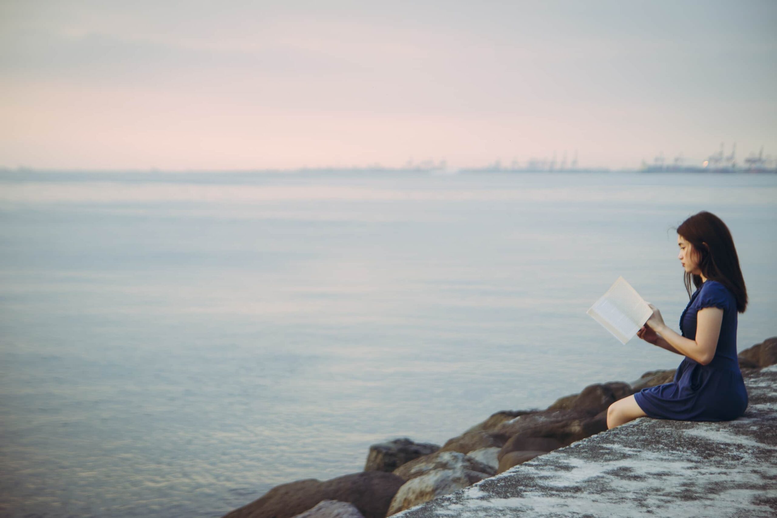 A woman is sitting on a sea wall reading a book. In the background is an ocean with a sunset.