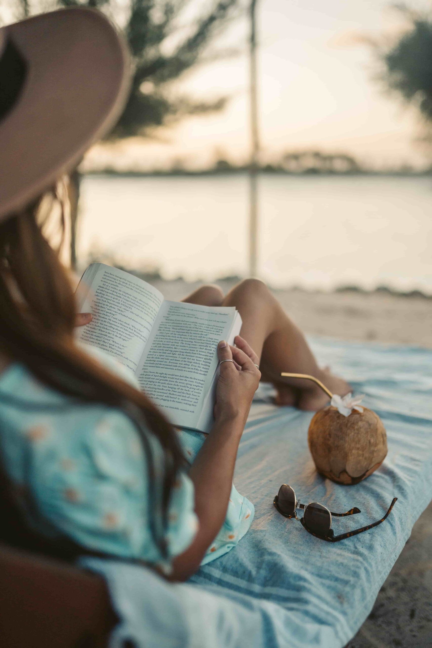 A woman is sat on a beach at sunset wearing a wide brimmed sun hat with an open book searched on her lap.
