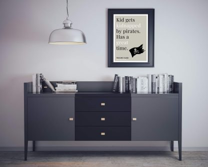 Humorous Treasure Island print in black frame on wall, with black sideboard and books below. From Bookish Prints collection by StrangeBooks UK