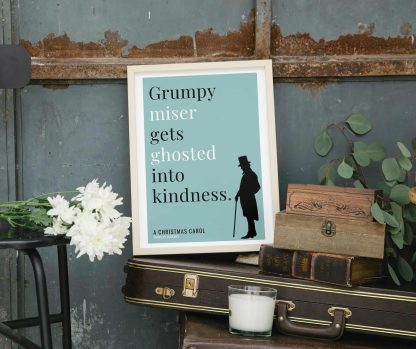Humorous Christmas Carol print in white wooden frame, perched on a violin case next to some books. From Bookish Prints collection by StrangeBooks UK