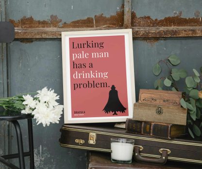 Humorous Dracula print in white wooden frame, perched on a violin case next to some books. From Bookish Prints collection by StrangeBooks UK