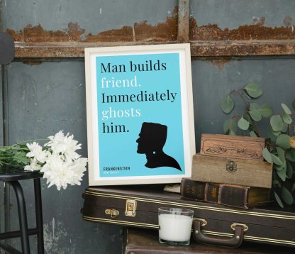 Humorous Frankenstein print in white wooden frame, perched on a violin case next to some books. From Bookish Prints collection by StrangeBooks UK