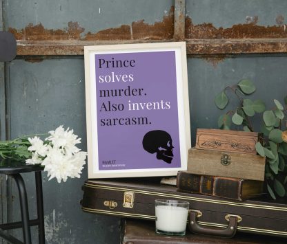 Humorous Hamlet print in white wooden frame, perched on a violin case next to some books. From Bookish Prints collection by StrangeBooks UK