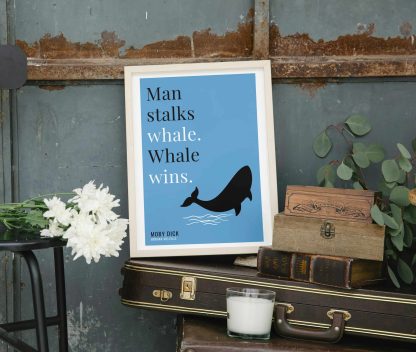 Humorous Moby Dick print in white wooden frame, perched on a violin case next to some books. From Bookish Prints collection by StrangeBooks UK