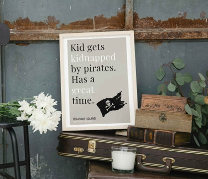 Humorous Treasure Island print in white wooden frame, perched on a violin case next to some books. From Bookish Prints collection by StrangeBooks UK