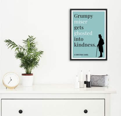 Humorous Christmas Carol print in black frame on wall, with white chest of drawers and plants and candles below. From Bookish Prints collection by StrangeBooks UK