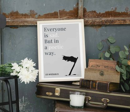 Humorous Les Misérables print in white wooden frame, perched on a violin case next to some books. From Bookish Prints collection by StrangeBooks UK