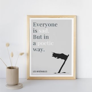 Humorous Les Misérables print in wooden frame, leaning against a plain wall. Small beige decoration nearby. From Bookish Prints collection by StrangeBooks UK