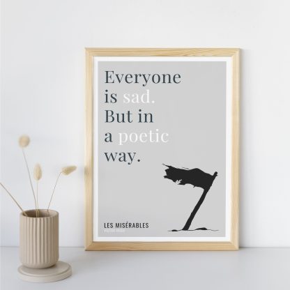 Humorous Les Misérables print in wooden frame, leaning against a plain wall. Small beige decoration nearby. From Bookish Prints collection by StrangeBooks UK