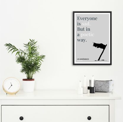 Humorous Les Misérables print in black frame on wall, with white chest of drawers and plants and candles below. From Bookish Prints collection by StrangeBooks UK