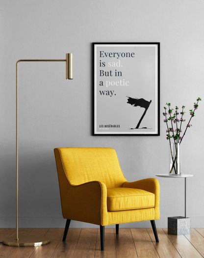 Humorous Les Misérables print in black frame on wall, with a yellow armchair in front next to a tall angular lamp and plant on a table. From Bookish Prints collection by StrangeBooks UK